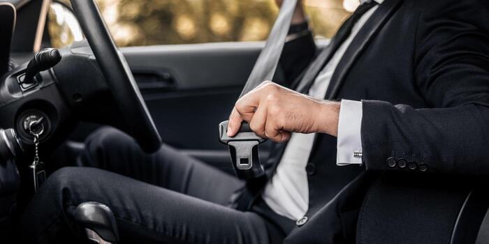 A driver in a suit reaching across to fasten the seat belt latch plate into the buckle beside the seat.